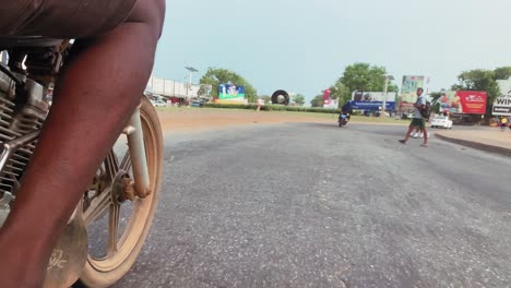 low-angle-point-of-view-shot-of-unrecognizable-motor-rider's-leg-and-front-tire-on-the-road-speeding-towards-a-roundabout-and-pedestrians-crossing-in-Tema,-Ghana
