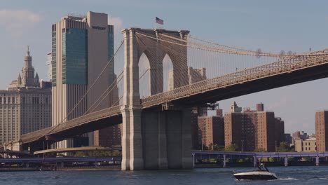 Wide-shot-of-beautiful-Brooklyn-bridge-with-motorboat-and-sailing-boat-cruising-below---New-York-City,USA