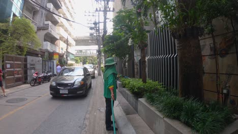 Female-gardener-watering-plants-with-hose-on-Bangkok-street-while-cars-drive-on-the-street