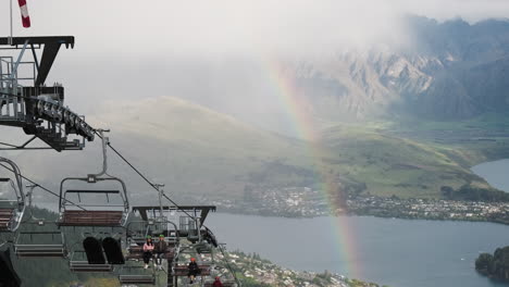 Rainbow-arcs-over-the-gondola-cars-as-they-pass-by-on-the-side-of-a-mountain-overlooking-Queenstown,-New-Zealand