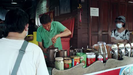 Mann-Macht-Tee-Und-Kaffee-In-Thailand,-In-Der-Nähe-Von-Bangkok-Auf-Dem-Schwimmenden-Markt-Damnoen-Saduak,-Während-Menschen-Herumlaufen-Und-Masken-Tragen