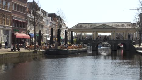 Overlooking-the-Nieuwe-Rijn-river-with-the-stone-arched-Kroomburg-bridge-in-the-distance,-as-people-go-about-their-day-enjoying-the-sunshine-at-a-coffee-shop-terrace-outside,-Leiden,-Netherlands