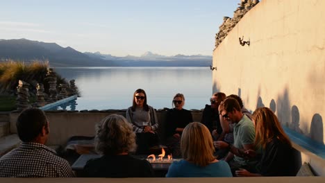 Friends-enjoying-drinks-around-fire-at-sunset-on-terrace-with-Mount-Cook-and-Lake-Pukaki-in-background