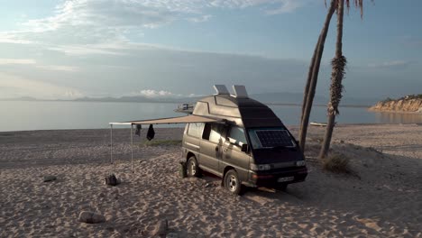 Camper-travel-van-with-side-canopy-extended-and-solar-panel-in-front-windshield,-Wide-handheld-shot