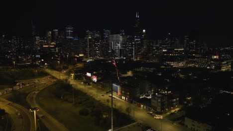 US-Flag-with-the-night-lit-Chicago-skyline-in-the-background---circling,-drone-shot