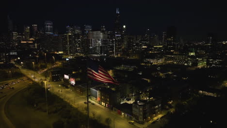 Aerial-view-passing-the-US-Flag,-tilting-toward-the-illuminated-skyline-of-Chicago-from-the-Fulton-River-district