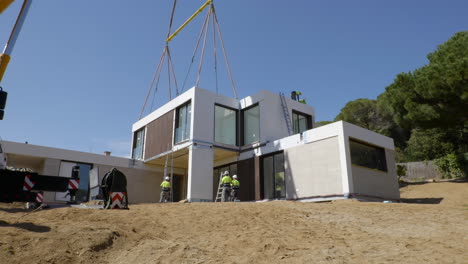 Construction-workers-installing-suspended-smart-modular-house-with-heavy-crane-lift-on-construction-site