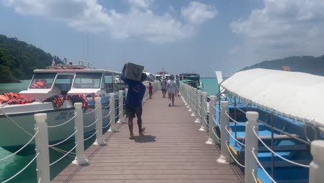 Man-carrying-cool-box-walks-along-jetty-surrounded-by-long-tail-boats