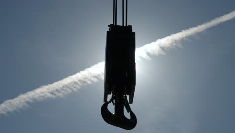 Silhouette-of-heavy-construction-crane-hook-and-tackle-swinging-against-sunlit-blue-sky-and-airplane-contrail-background