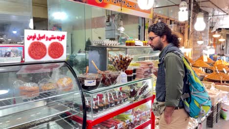 Ein-Mann-Kauft-Eine-Keksdose-In-Einer-örtlichen-Konditorei-Auf-Dem-Bauernmarkt-Im-Iran-Und-Wartet-Auf-Süße-Und-Frisch-Gebackene-Kekse.-Der-Kuchen-Wird-Einer-Ladenbesitzerin-Bezahlt,-Die-Im-Laden-Mit-Traditionellen-Produkten-Arbeitet