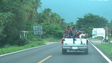 Three-men-riding-on-the-back-of-a-pickup-truck-full-of-cargo-on-a-two-lane-road-near-the-coast,-Dashboard-view
