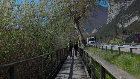 Back-view-of-couple-of-hikers-walking-along-wooden-catwalk-near-busy-road,-Toblino-Lake-in-Trentino,-Italy