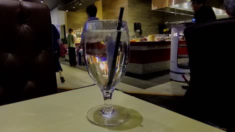 closeup-of-clear-glass-cup-with-water-and-black-straw-in-the-foreground-while-background-AI-robot-directs-people-to-seating-area-at-fast-food-eatery-buffet-all-you-can-eat-multicultural-people-place