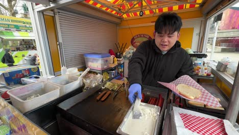 Famous-Seattle-style-hot-dog-from-Asian-male-vendor-at-food-cart-preparing-on-grill-with-onions,-peppers-and-cream-cheese