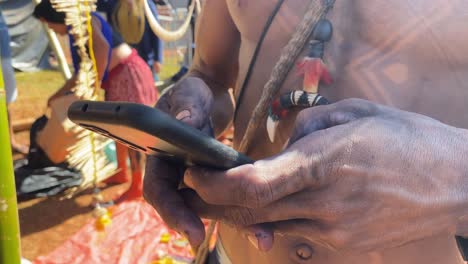 Eine-Nahaufnahme-Eines-Farbenfrohen,-Traditionell-Bemalten-Und-Mit-Federhut-Gekleideten-Einheimischen-Amazonas-Mannes,-Der-Auf-Sein-Smartphone-Schaut