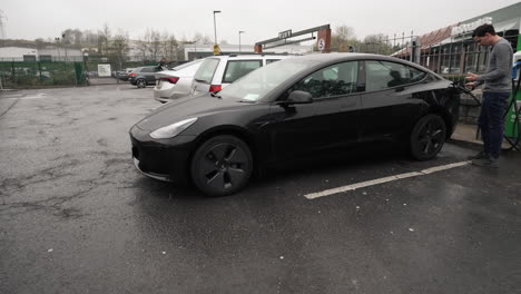Panning-shot-of-a-young-man-charging-a-Black-Tesla-EV-on-a-rainy-day-in-Europe