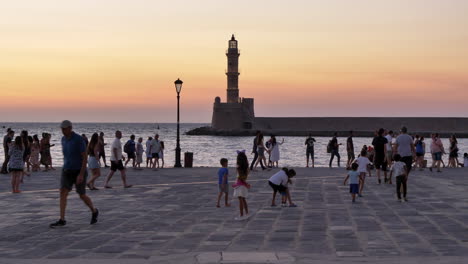 People-on-promenade-at-sunset,-lighthouse-in-background,-Chania