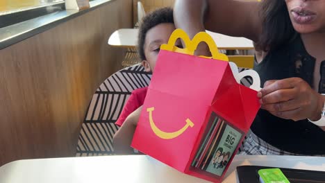 Surprised-3-year-old-black-boy-opening-a-Mc-Donald’s-Happy-Meal,-sitting-next-to-his-beautiful-young-balck-mother