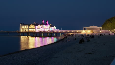 Nächtlicher-Zeitraffer-Der-Sellin-Pier,-Beleuchtet-Mit-Farbenfroher-Beleuchtung-Während-Des-Konzerts,-Silhouette-Von-Menschen,-Die-Am-Kiesstrand-Der-Insel-Rügen-An-Der-Ostsee-Spazieren