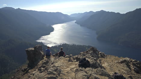 Two-young-men-sitting-on-scenic-ridge-overlooking-beautiful-mountain-lake