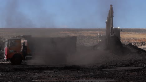 Excavator-load-crushed-coal-into-dump-truck,-dusty-mine-site-environment