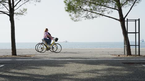 Menschen,-Die-An-Der-Strandpromenade-Von-Thessaloniki-Vorbeikommen-Und-Fahrräder-Und-Elektrofahrräder-Fahren,-Meereshintergrund,-Sonniger-Tag-Im-Sommer