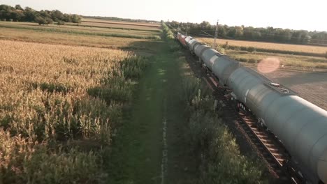 Aerial-drone-footage-closely-follows-a-loaded-cargo-tank-train-through-vast-golden-lit-cornfields,-the-drone-flying-just-beside-the-wagons,-revealing-the-serene-rural-landscape