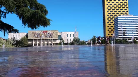 People-cool-off-in-front-of-the-fountains-in-the-center-of-the-capital-Tirana-in-hot-summer-days,-National-Museum-and-high-buildings-city-background
