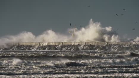 Wide-shot-of-a-giant-waves-crashing-over-the-breakwater-with-a-navigation-beacon-in-the-background-as-seagulls-fly-over-the-water,-slow-motion