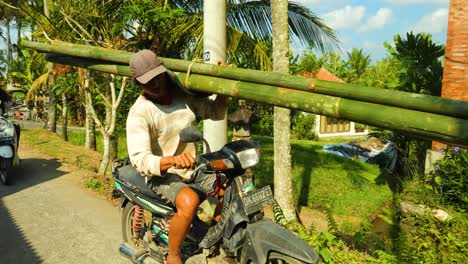 Zeitlupenaufnahme-Eines-Fahrenden-Bauarbeiters-Auf-Einem-Roller-Mit-Bambusstöcken-Auf-Der-Schulter,-Der-Zur-Baustelle-Auf-Bali,-Indonesien-Fährt