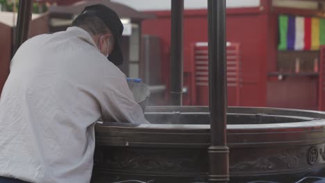 Man-Removing-Ash-From-Incense-Burner-At-Senso-ji-Temple-In-Asakusa,-Tokyo,-Japan