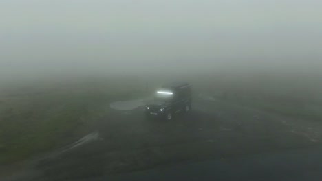 Drone-tilt-up-of-black-Land-Rover-Defender-parked-in-very-cloudy-and-misty-mountain-hills-with-zero-visibility-and-fog-lights-shining