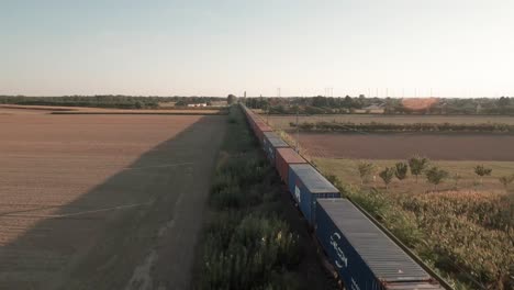 Beladene-Reise:-Luftaufnahmen-Verfolgen-Einen-Beladenen-Güterzug-Mit-Containern-Durch-Weite,-Goldbeleuchtete-Maisfelder
