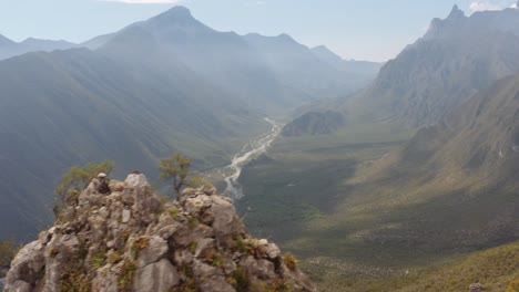 Drone-flying-past-rocky-mountain-peak-pans-left-to-reveal-dam-in-valley-far-below