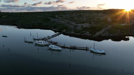 Sun-flare-glistens-above-ridgeline-on-natural-port-dock-of-Sa-Nitja-Menorca-Spain,-calm-water-reflects-cloudy-sky