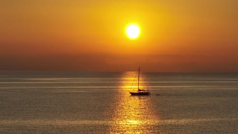 Side-view-of-backlit-sailboat-silhouette-floating-on-open-ocean-in-sun-ray-beam-under-orange-golden-hour-sky