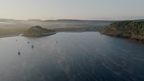 Meeresnebel-Erhebt-Sich-In-Mystischen-Rankenartigen-Wellen-über-Boote,-Die-Am-Unberührten-Strand-Von-La-Vall-Menorca-Vor-Anker-Liegen