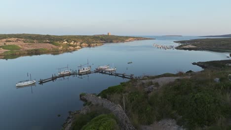 Drone-descends-along-rock-wall-to-wooden-harbor-dock-of-fishing-boats-in-Sa-Nitja-Menorca-Spain