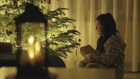 Ein-Mädchen-Sitzt-Neben-Dem-Weihnachtsbaum,-Hält-Ein-Geschenk-In-Der-Hand-Und-Berührt-Sanft-Die-Hängenden-Dekorationen-–-Nahaufnahme