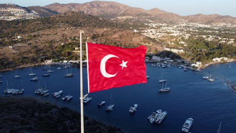 Aerial-static-shot-of-Turkey-flag-fluttering-in-wind,-Gumusluk-coast-in-background