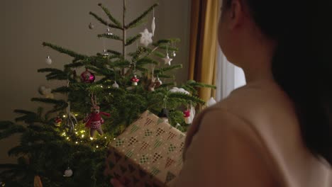 Ein-Mädchen,-Das-Ein-Geschenk-In-Der-Hand-Hält,-Untersucht-Die-Dekorationen,-Die-Am-Weihnachtsbaum-Hängen-–-Mittlere-Nahaufnahme
