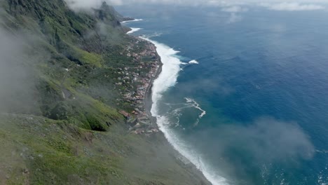 Misty-thin-wispy-clouds-hang-low-on-green-mountains-towering-above-coastal-town-in-Madeira-Portugal