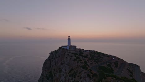 Aerial-pullback-as-dusk-sky-surrounds-Formentor-lighthouse,-Serra-de-Tramuntana-Mallorca-Spain-as-car-drives-up