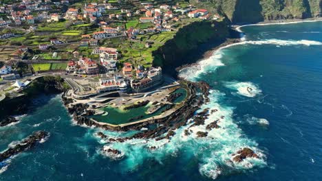 Drone-orbit-around-natural-pools-and-town-of-Porto-Moniz,-Madeira-Portugal-coastline-on-sunny-day