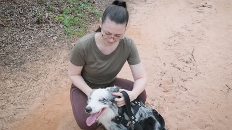Woman-with-glasses-having-a-nice-time-with-her-pet-Australian-Shepherd-dog,-petting-him-and-playing-with-him-in-the-forest