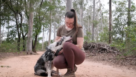 Woman-with-glasses-having-a-nice-time-with-her-pet-in-a-forest,-Australian-shepherd-dog,-caresses-him-and-kisses-him