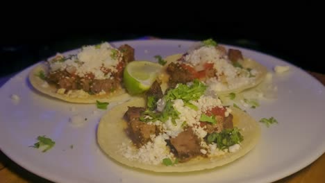 3-meat-tacos-optical-zoom-closeup-unfolded-corn-tortilla-topped-green-onions-parsley-lime-sliver-salsa-on-white-plate-on-top-of-wood-table-with-dark-background-movement