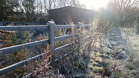 Frost-covered-wooden-fence-surrounding-frozen-farmland-barn-building-during-golden-hour
