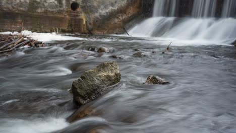Winter-river-flowing-over-rocks-below-dam-sluice-waterfall,-timelapse