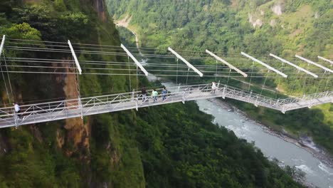 Aerial-view-of-people-crossing-suspension-bridge-over-river-in-green-mountains-of-Nepal